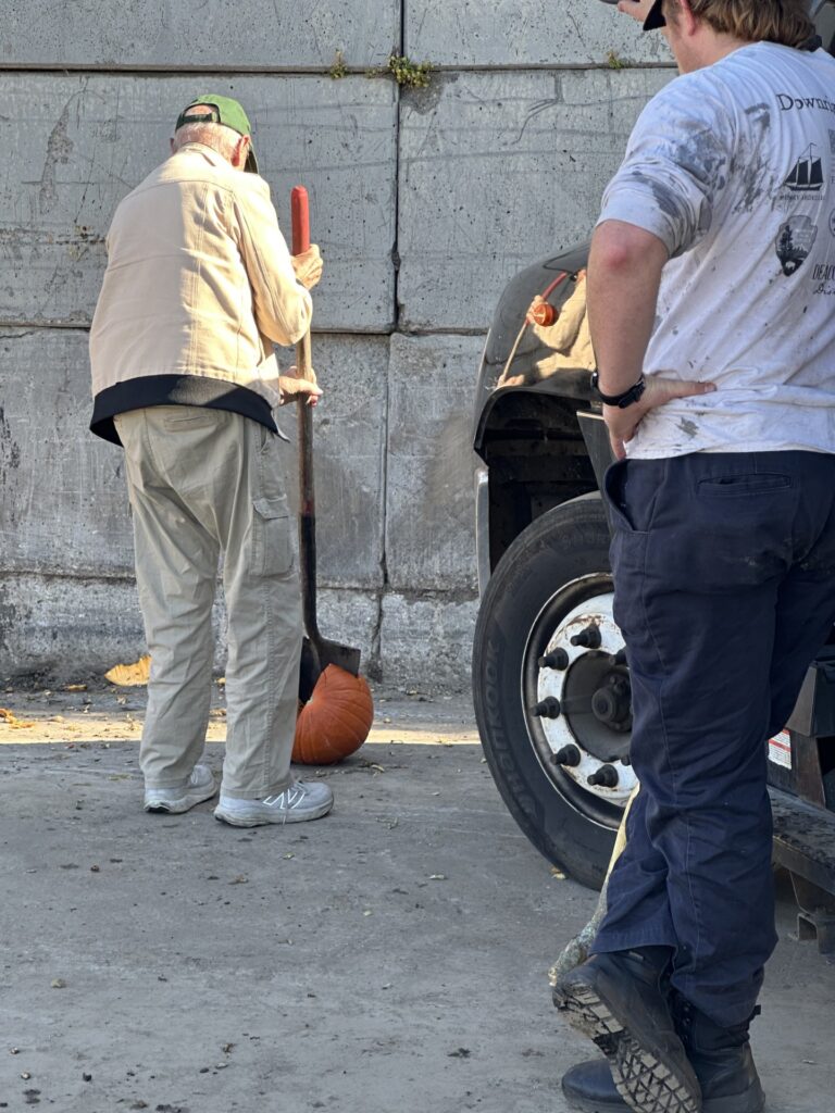 A man, shown from the back, takes a shovel to a pumpkin. Someone else watches from the side.