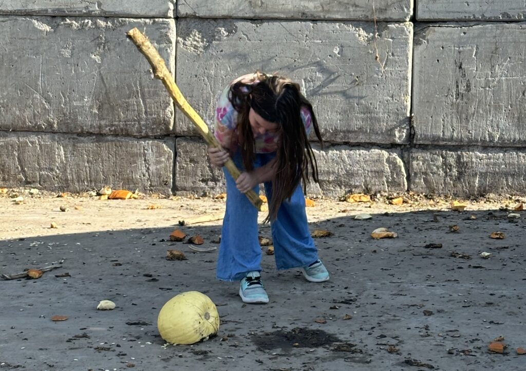 A young girl speaks softly but takes a big stick to a large pumpkin.