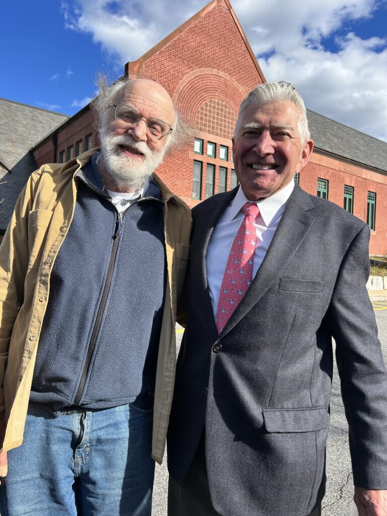 Two men stand with a brick facade in the background.