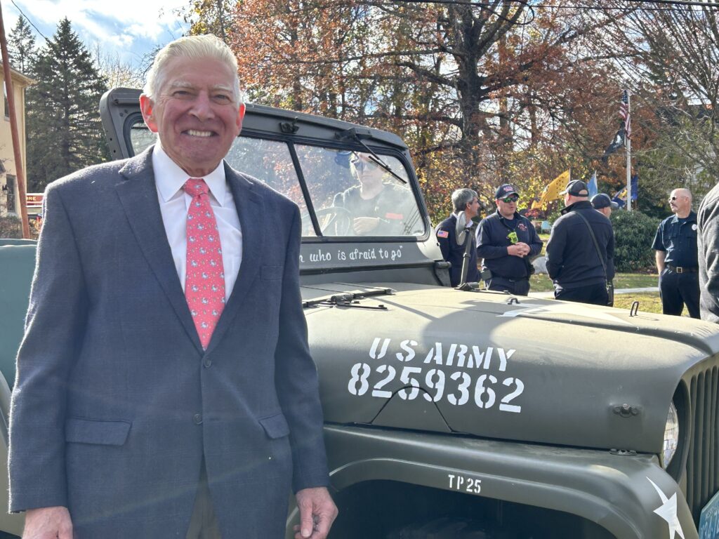 An older man in a gray suit and red tie smiles in front of an Army-issue green Jeep.