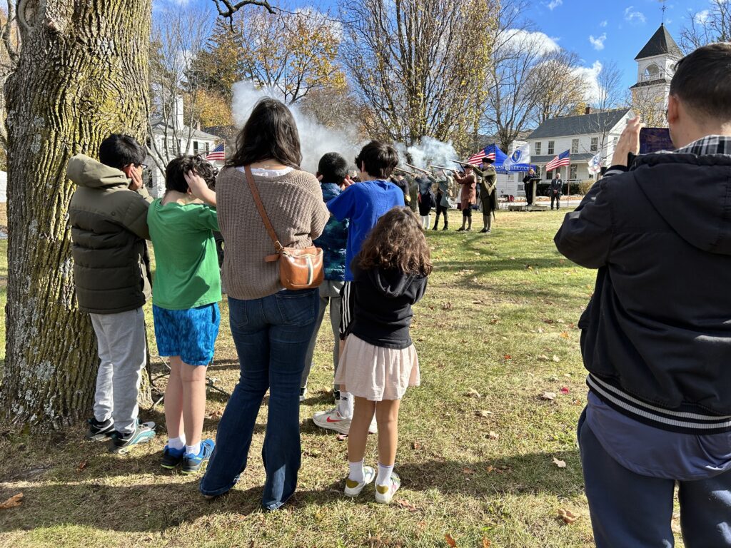 A group of people on the left watch the Minutemen shoot their muskets. The kids have their hands over their ears as smoke drifts away from the guns in the background.