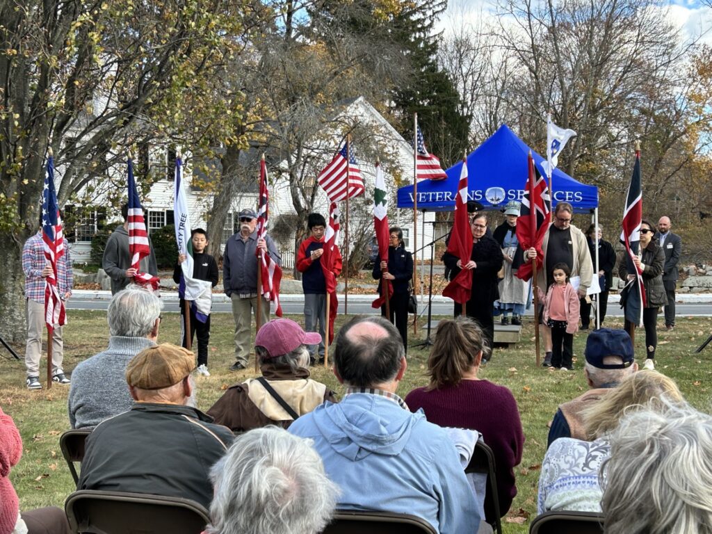 Several people lined up holding flags.There are people seated in the foreground and the Veteran's Services tent in the background.