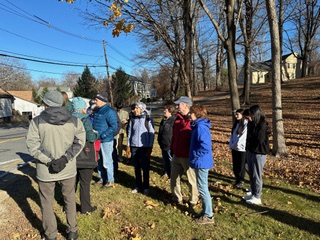 A group of people wearing outdoor clothing stands in the sun outside. A yellow building is visible at the top of a hill on the right side of the image.