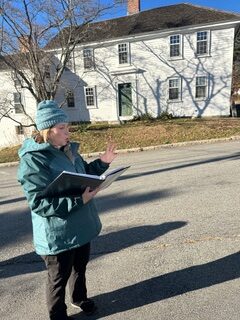 A woman in a blue coat and holding a large notebook, speaks in front of an old white building.