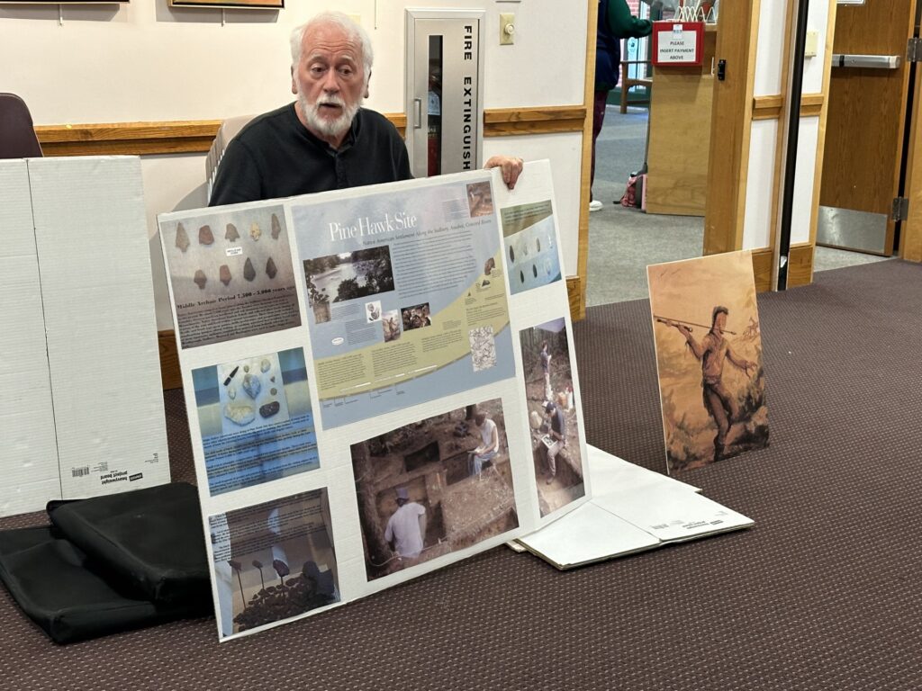 A bearded man holds a large tri-fold poster that contains information about the Pine Hawk archeological site.