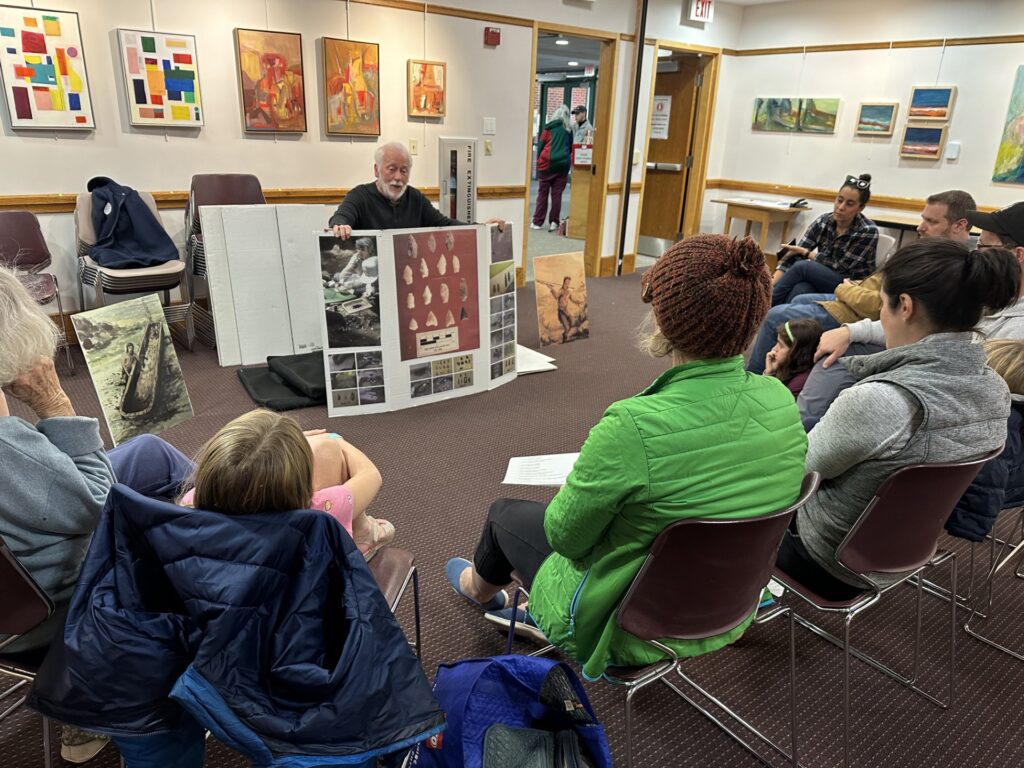 An older man sits in front of an audience holding a large tri-fold poster. the center of the poster has what looks like pictures of arrowheads and there are a number of other pictures on the poster.