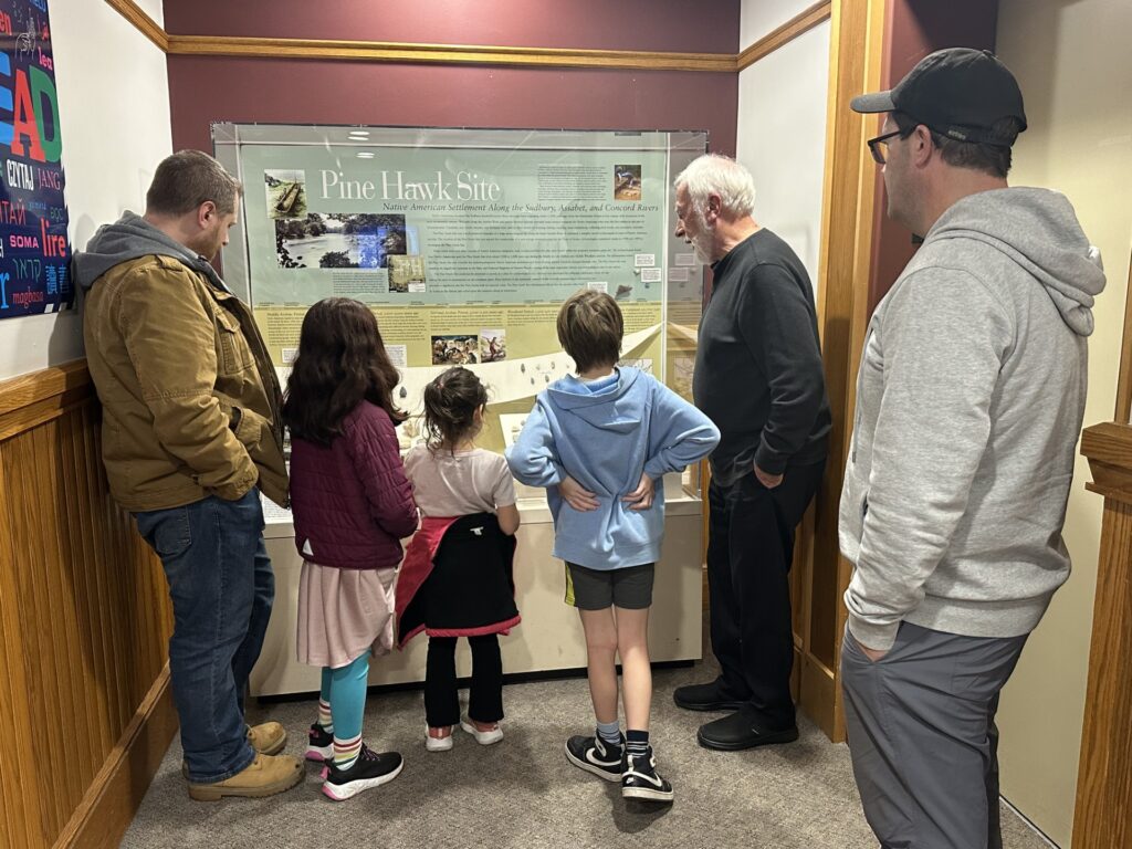 Three kids and a couple of parents stand in front of a glass fronted exhibit. A bearded older man is on the side describing the objects within.