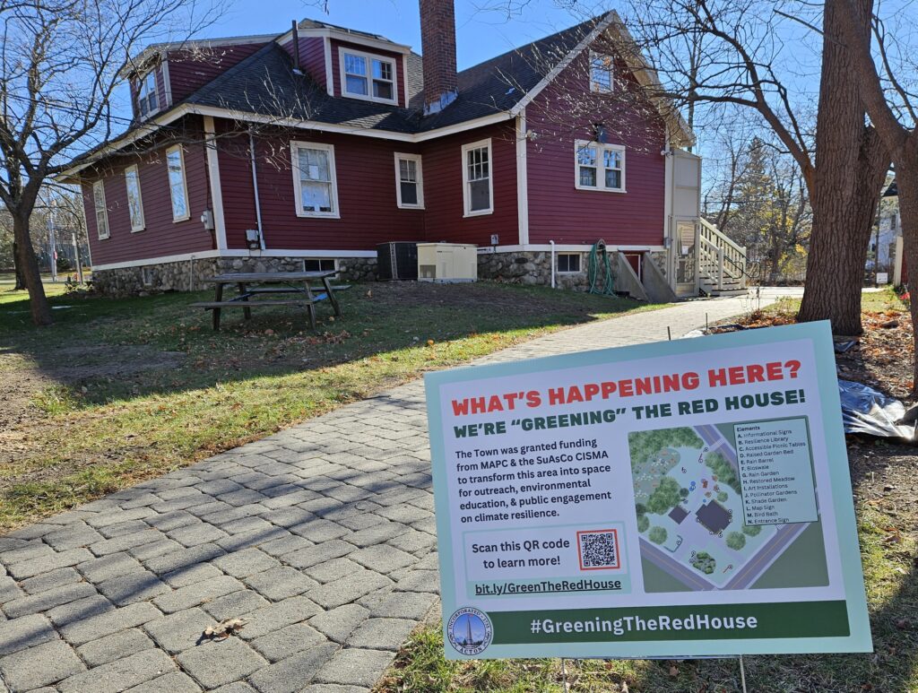 An older red house in the sun. In the foreground, a yard sign says: What's happening here? We're "Greening" the Red House Along with additional information.