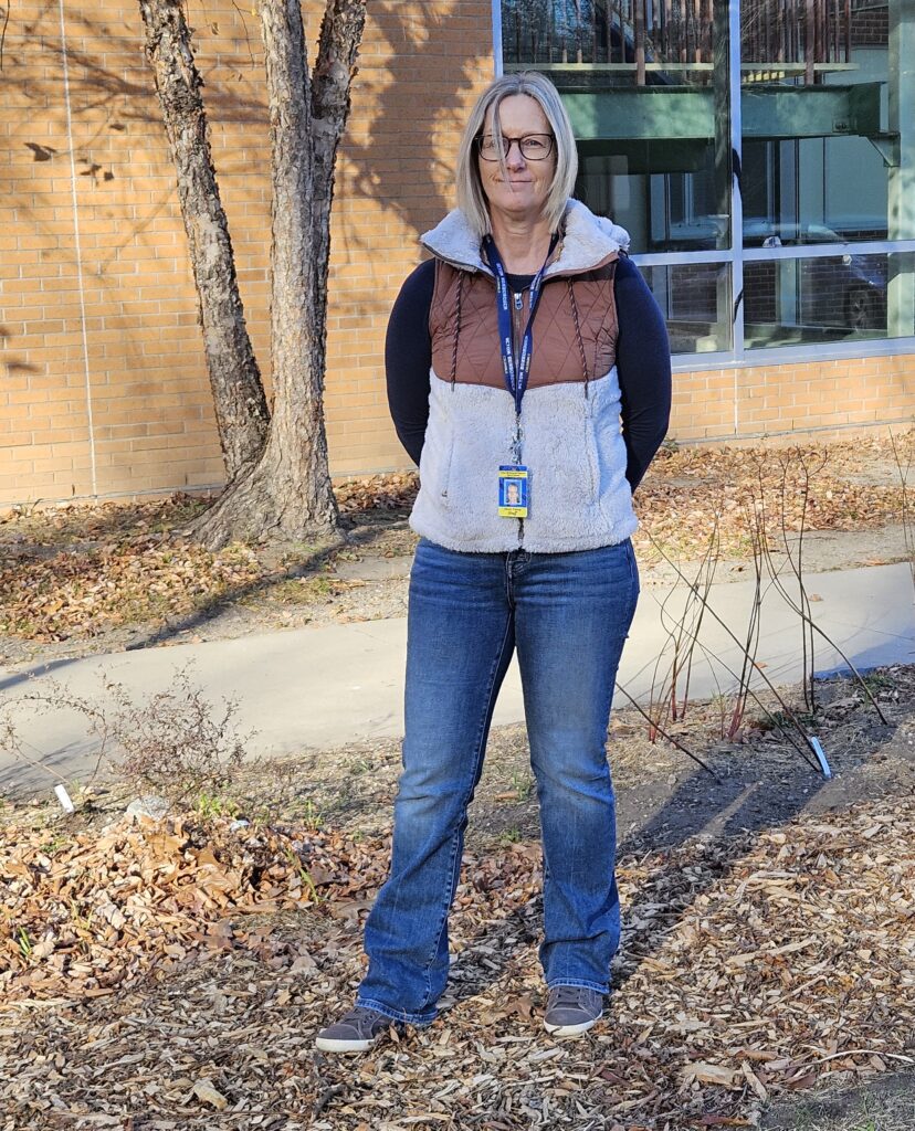 A woman with blonde hair wearing a vest and jeans stands in front of a wall at the high school.