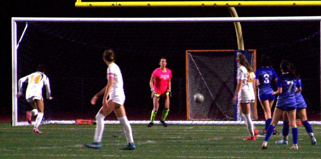 A group of young women in soccer gear are in front of the goal. The ball is heading towards the goal, and the goalie is heading towards the ball.