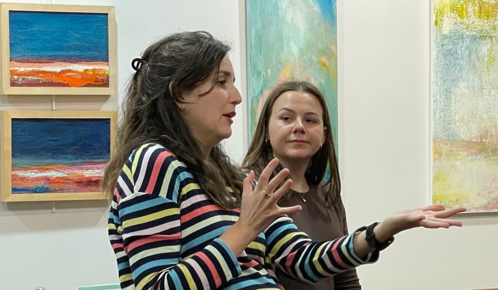 Two women. The woman in the foreground is speaking and using her hands to gesture. The woman in the background is watching her. On the wall, art from this month's AML exhibition can be seen.