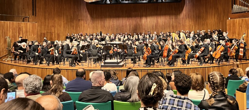 A large stage full of classical musicians and a chorus behind them. The stage is about as full as it can be.