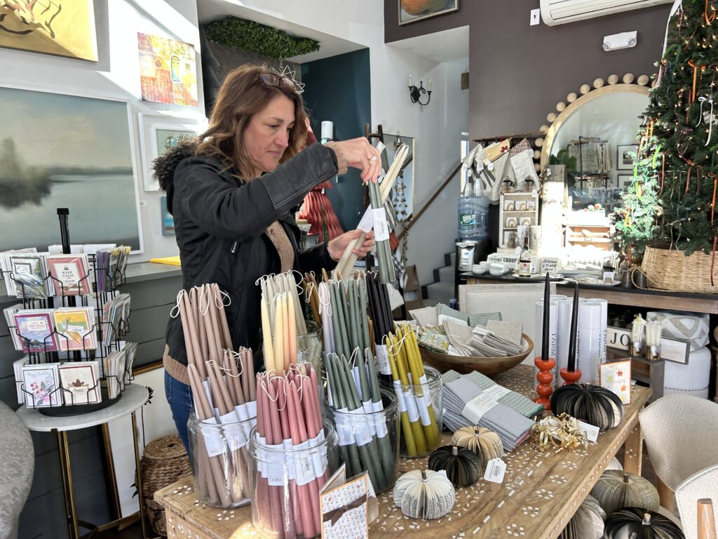 Woman with long brown hair aranges a table full of colorful candles. The table contains other things as well and there is a small card rack on her left.