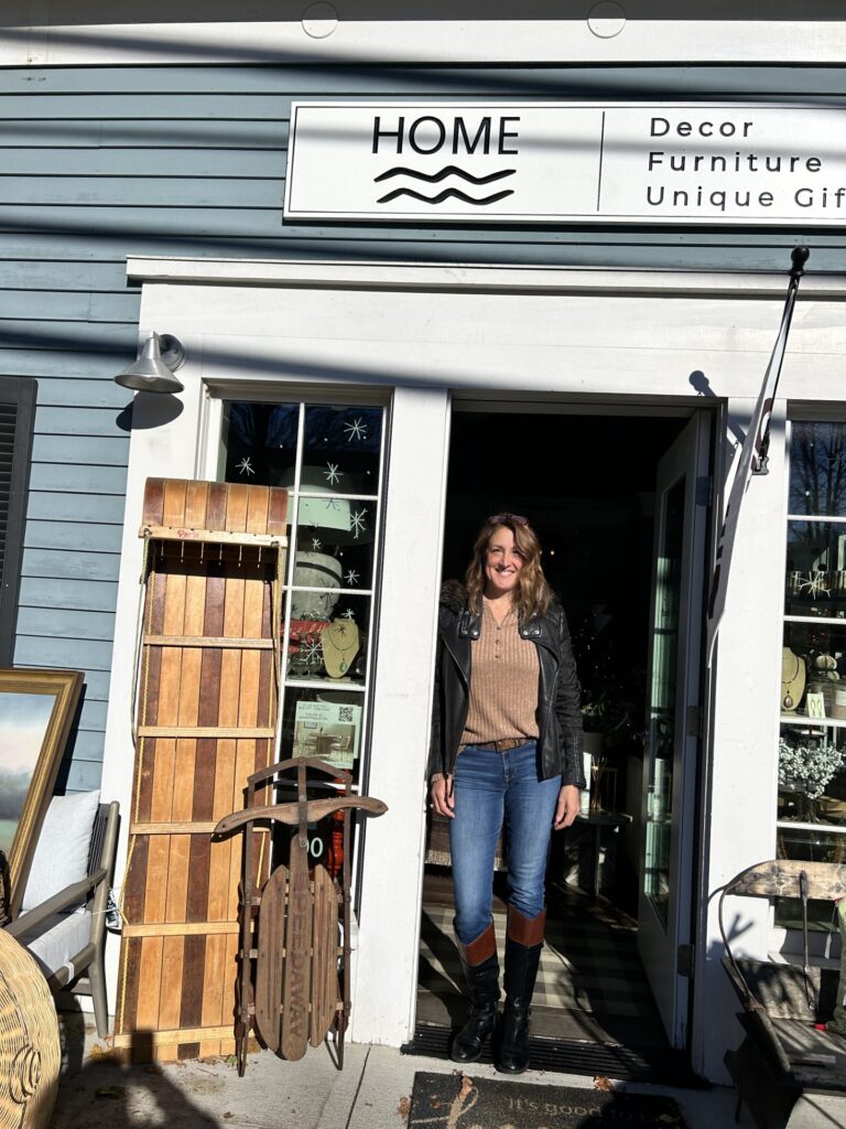 A woman stands in a sunny doorway. A sign above the door Reads HOME: Decor, Furniture, Unique Gifts, and miscellaneous items decorate the doorway, including two old sleds.