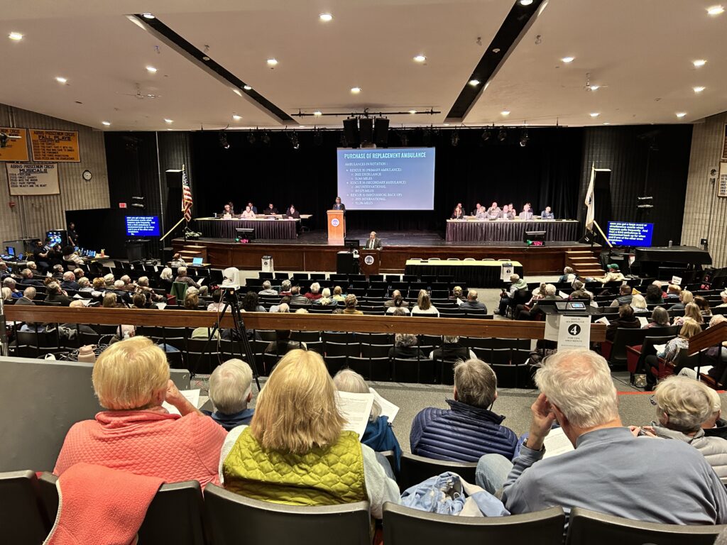 View of stage, very far and small, with people seated at long tables to the elft and right of a screen showing a slideshow. Closer are rows of audience members from behind, not filling up every seat, and closest are four people just close to the camera, also from behind, two blonde women wearing orange and green, a man with grey hair and a grey shirt.