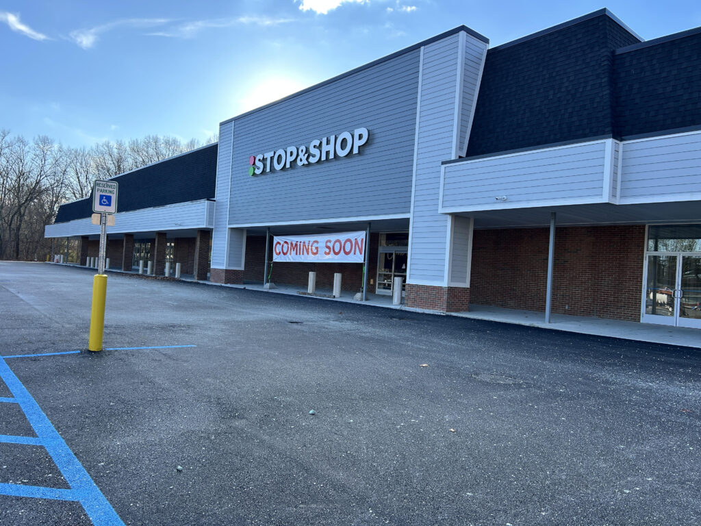 Handicapped parking sign in foreground, with blue painted lines on the pavemnt. Diagonal view of a long, low building facade, light grey horizontal wood on top, brick on bottom, with large signs: "Stop & Shop", and "Coming Soon".