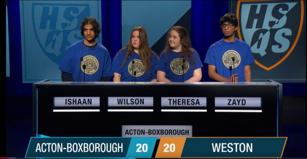 Four kids; two boys, two girls, wearing blue and yellow AB t-shirts stand behind a game show podium. A sign that says HSQS is in the background.