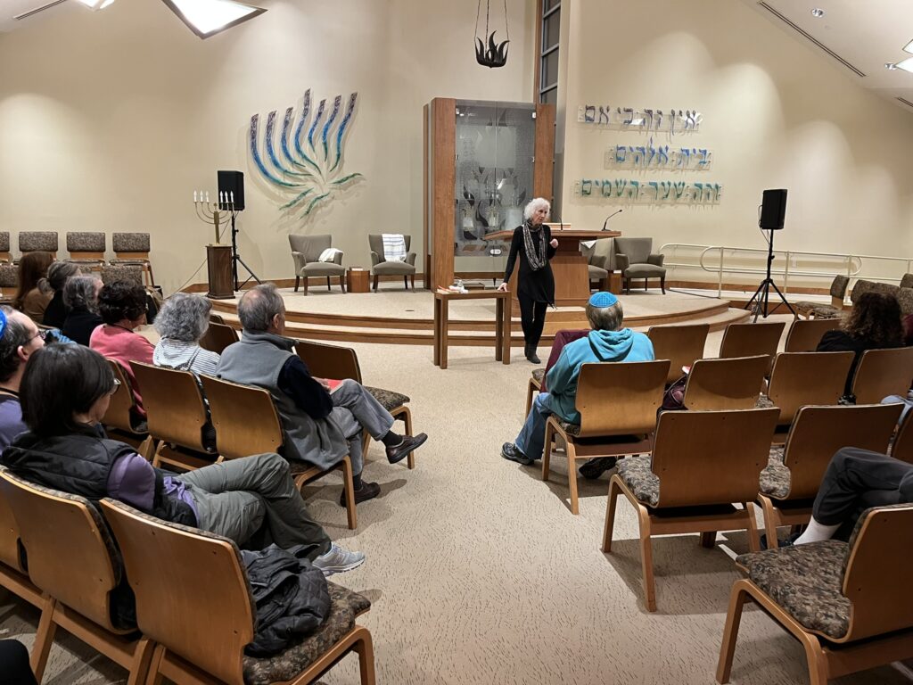 A silver-haired woman, elegently dressed in black with a colorful scarf, stands at a table in a synagogue sanctuary. A number of people sit in the audience. The sanctuary has an artistic menorah on the left side of the wall and Hebrew writing on the right.