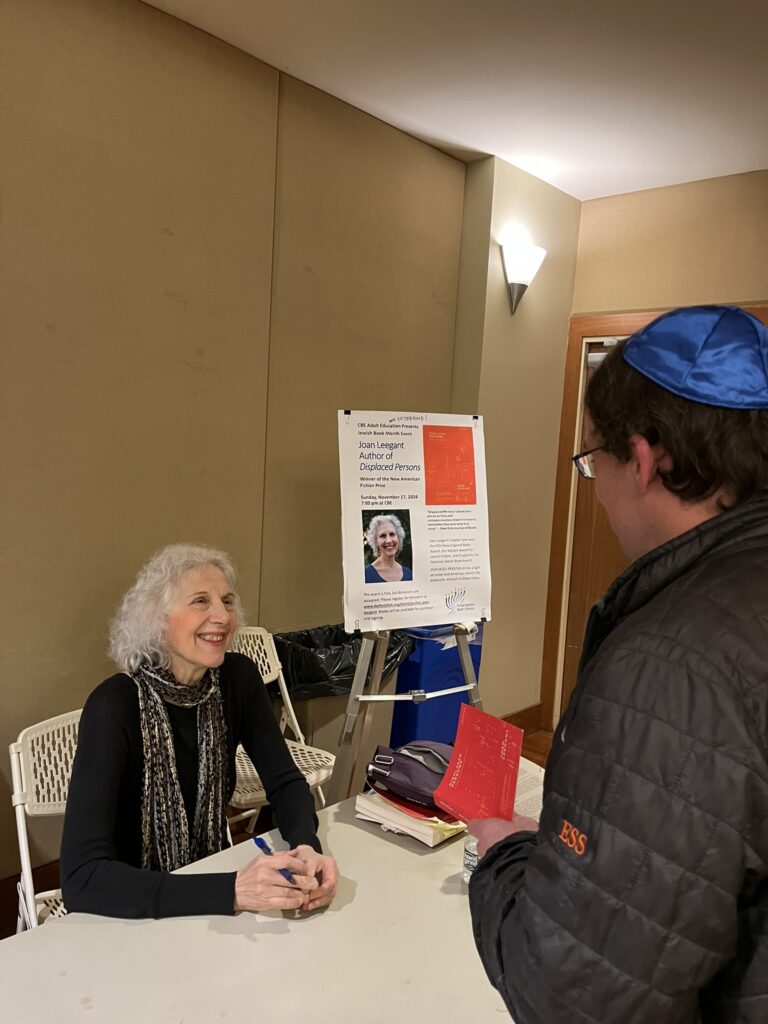 A smiling silver-haired woman sits at a table speaking to a man wearing a kippah. She is holding a pen, he is holding a book. It seems that that the book will be signed soon.