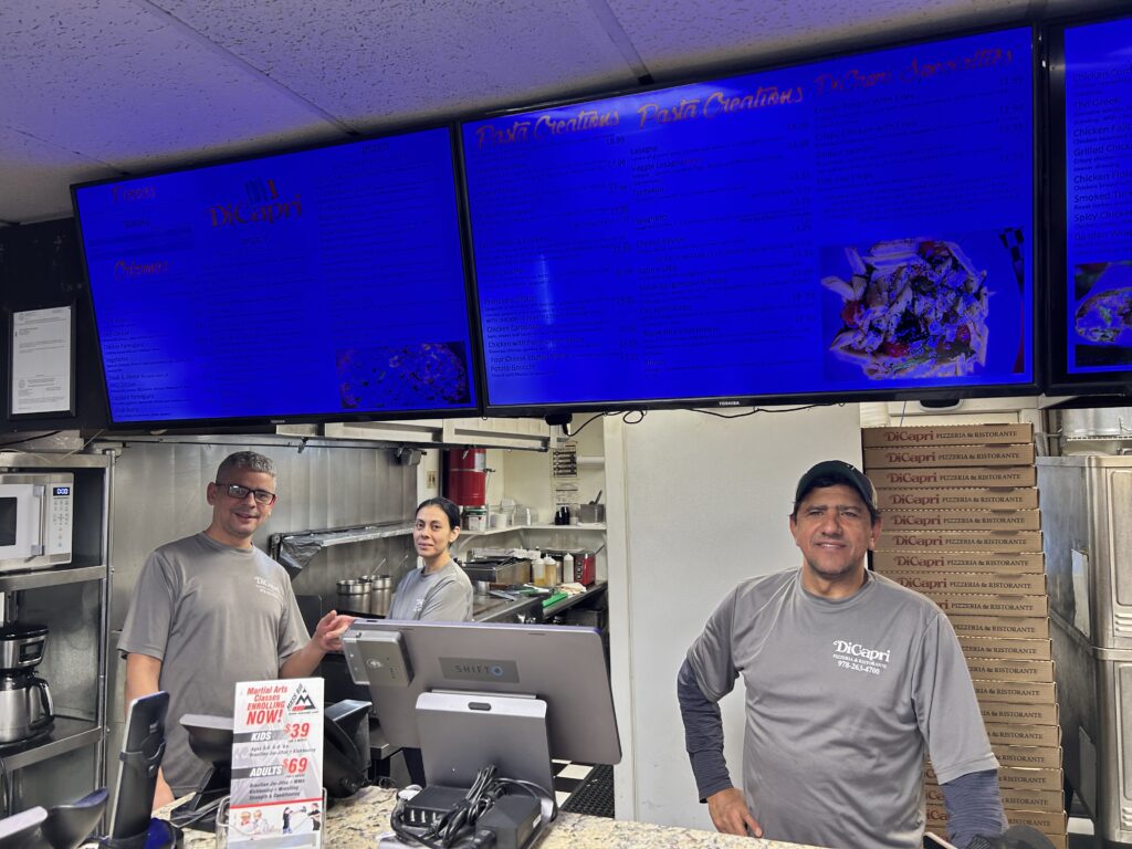 Three people in gray t-shirts behind the counter at a pizza shop.