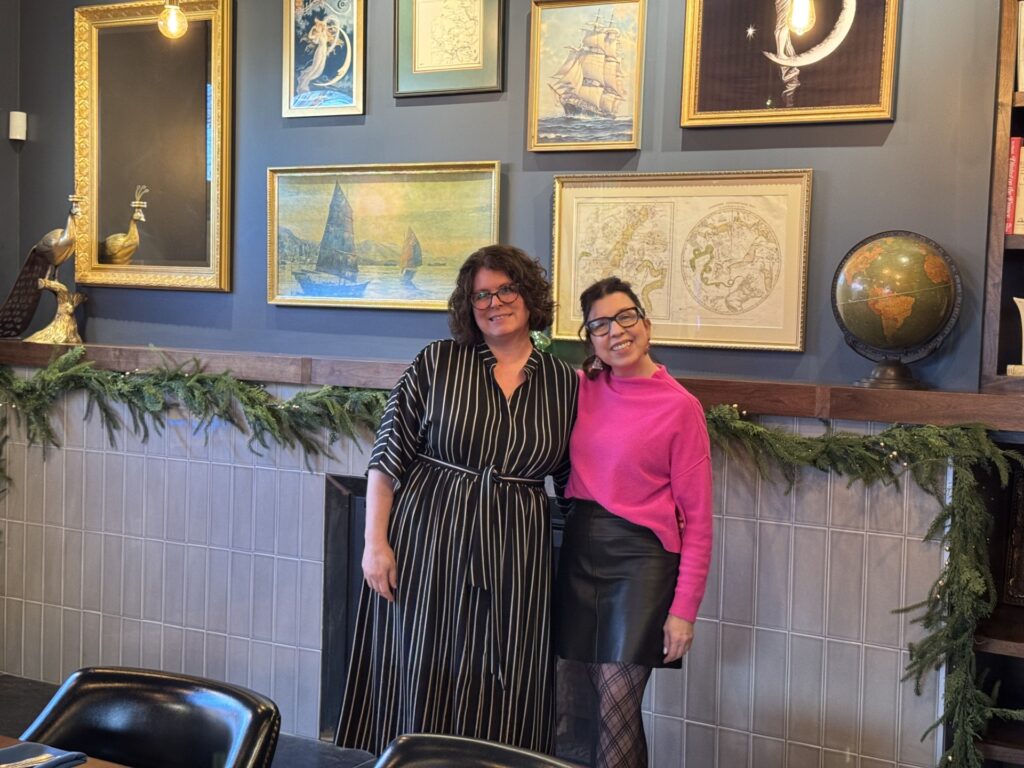 Two women stand in front of a mantel decorated with greens. The walls are blue and covered with pictures with nautical and lunar themes.