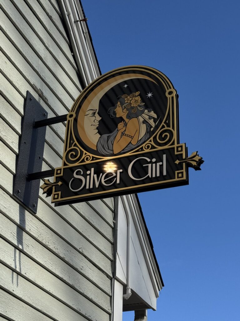 An Art Deco sign of a woman holding on to a smiling crescent moon. The sign stands out from the wall in front of a clear blue sky.