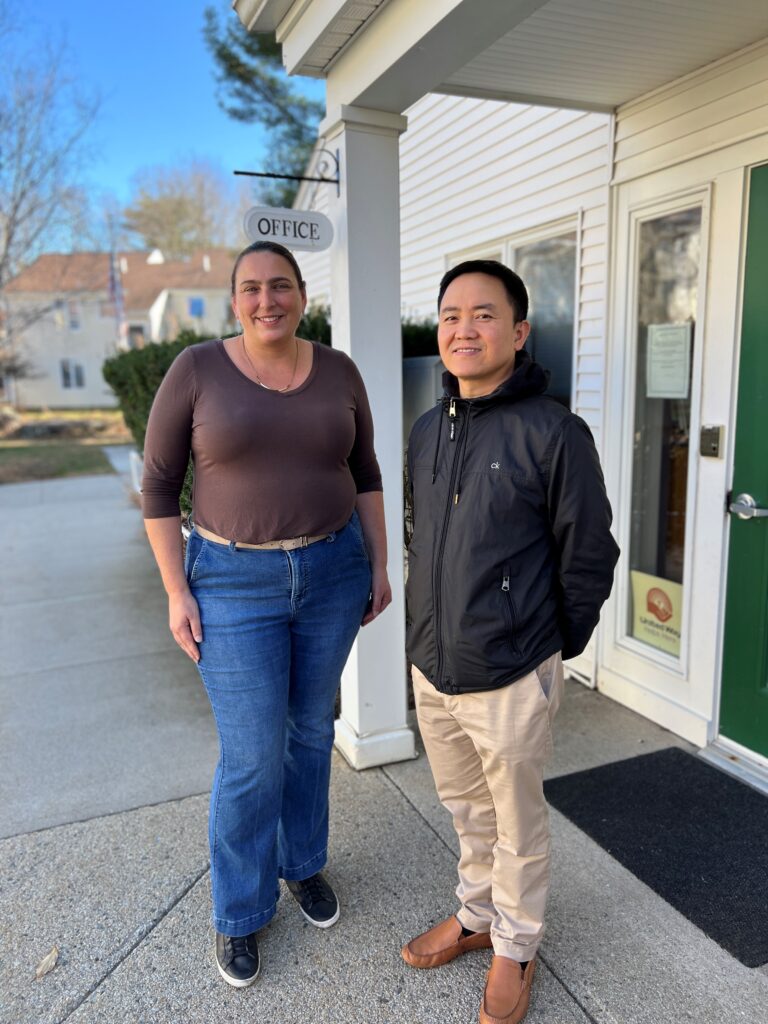 A tall woman and less tall man stand in front of a building. Behind them, a little Office sign lets people know that they are at the right place.