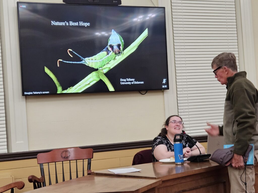 A woman sits at a desk talking to a standing man. Behind her, a slide on a screen says "Nature's Best Hope" and has a very large picture of an green insect on a stick.