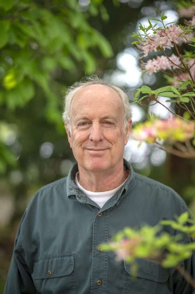 A balding older man stands by a flowering fruit tree.