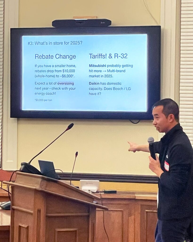 A man stands at a podium. Behind him, a screen describes some of the electrification rebate changes for 2025 as well as possible tariffs.