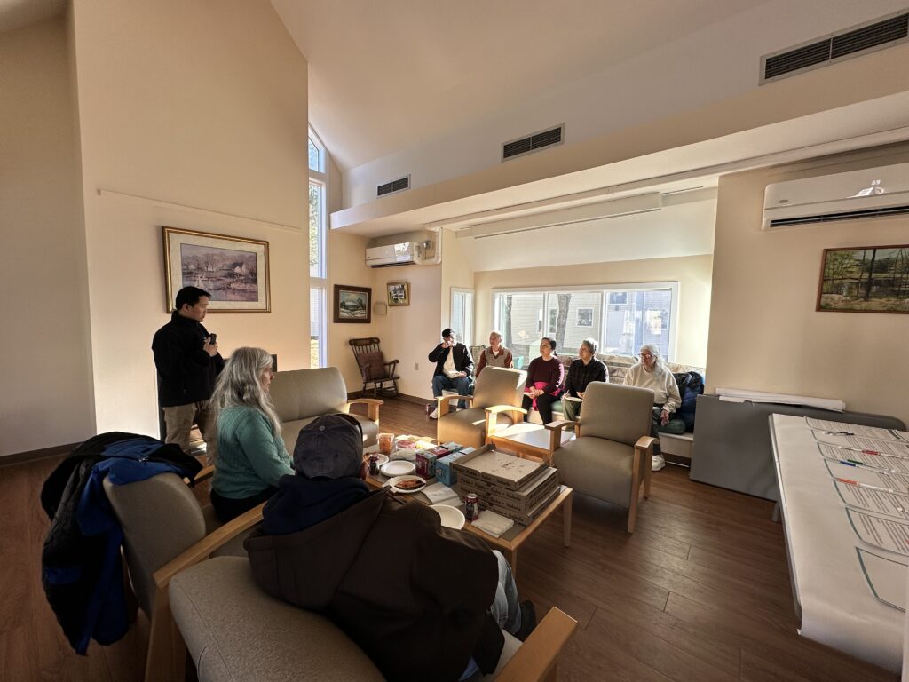 A bunch of people sit in a sunny room. There are three people on once side and five on the other, and a table with pizza and drinks in between.