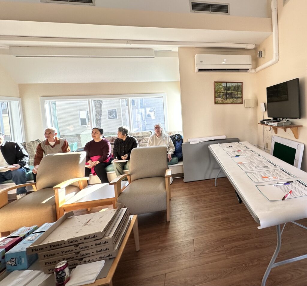 On the right, a table with papers laid out and multi-colored pens. On the back, a number of people sitting on a couch, listening, and in the foreground, pizza boxes.