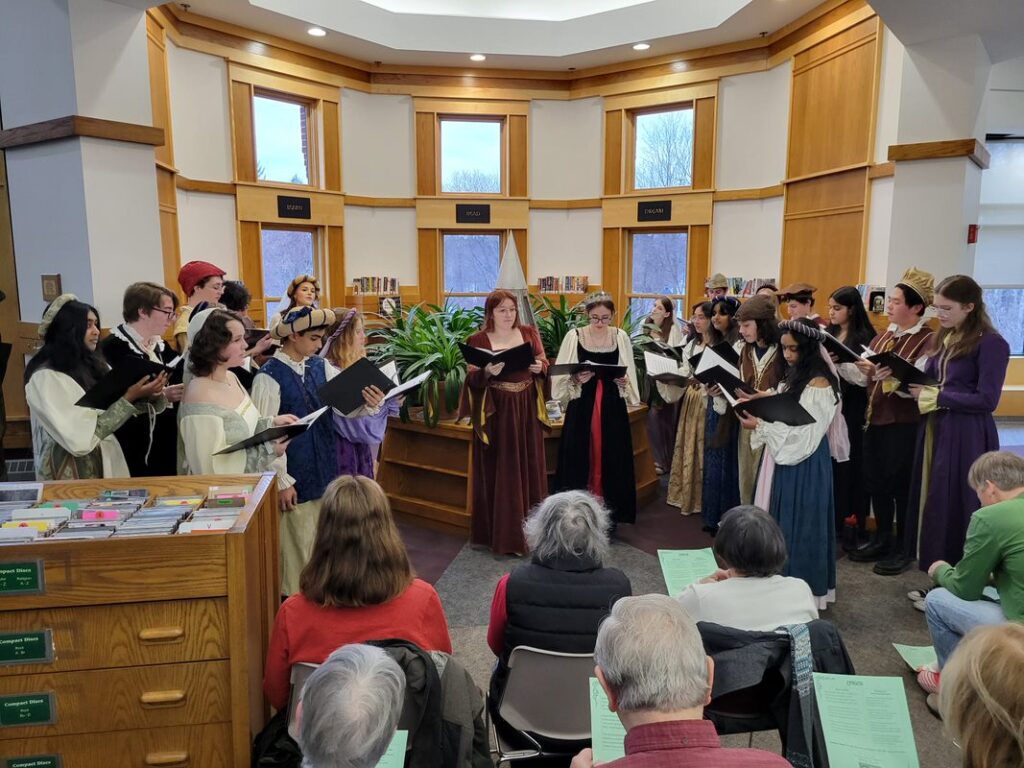 A large group of students in Renaissance dress stand in front of windows holding large books of music.