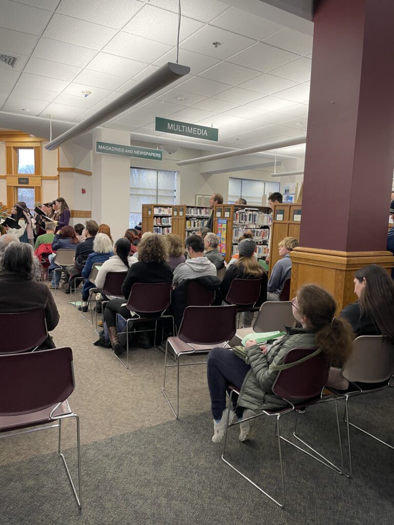 Rows of people seated with the madrigal singers in the background. A number of people are standing and watching from the library stacks.