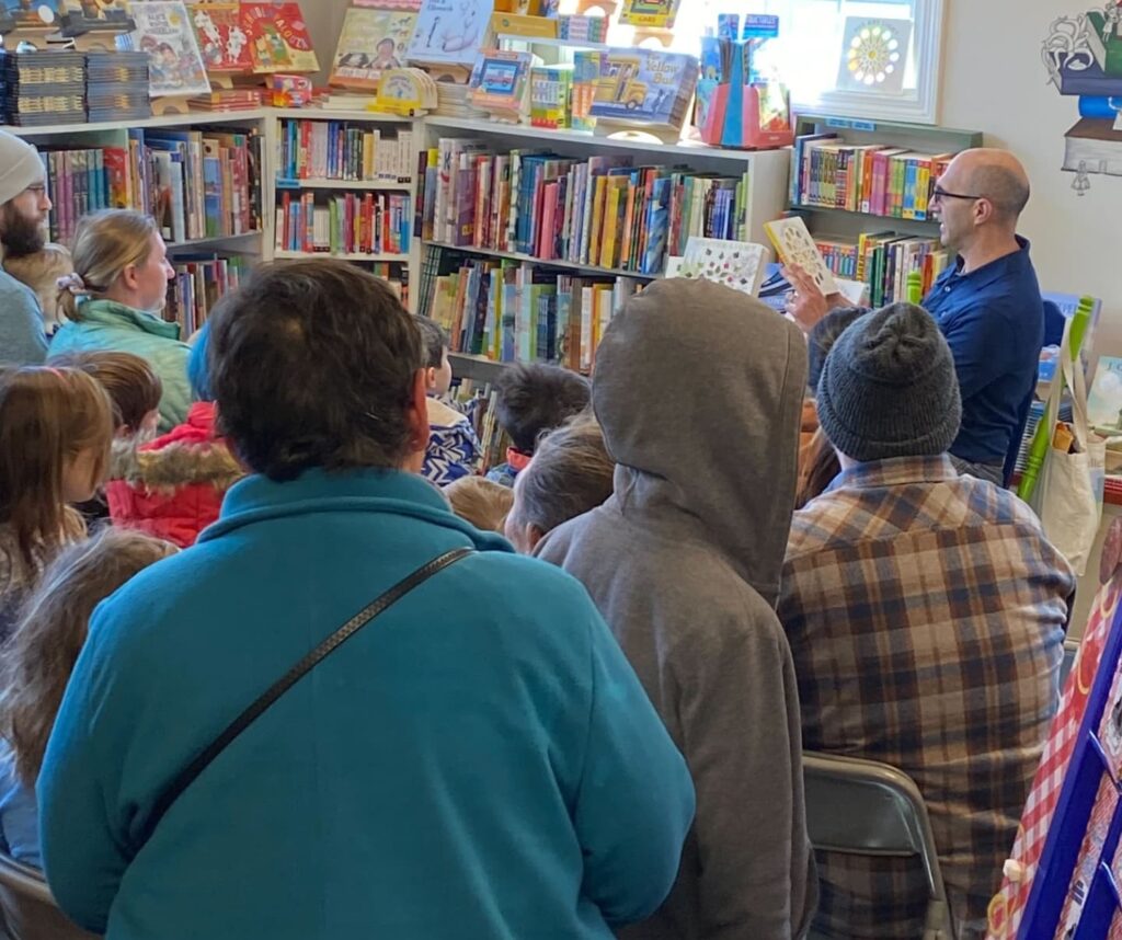 A man sits in a green chair holding up a book in front of a group of kids and adults.