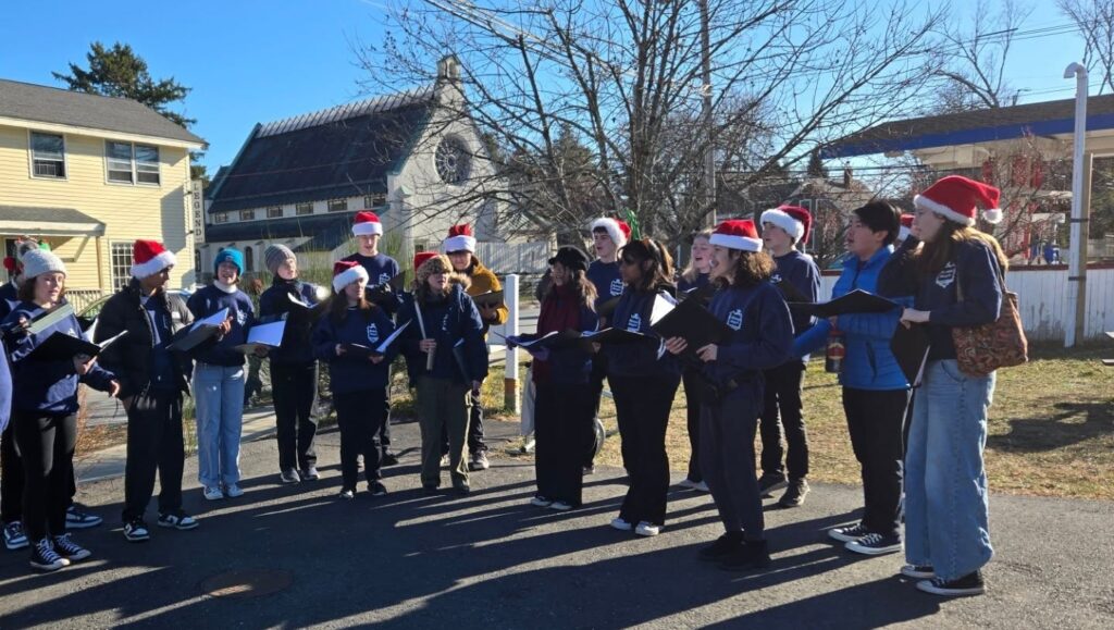 A group of teens wearing matching blue sweatshirts hold notebooks and look ready to sing. Many of the kids are wearing Santa hats.