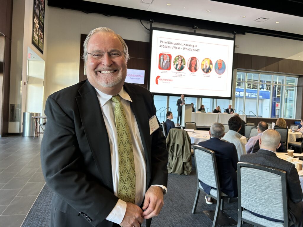 A man stands in a room full of people watching a panel discussion.