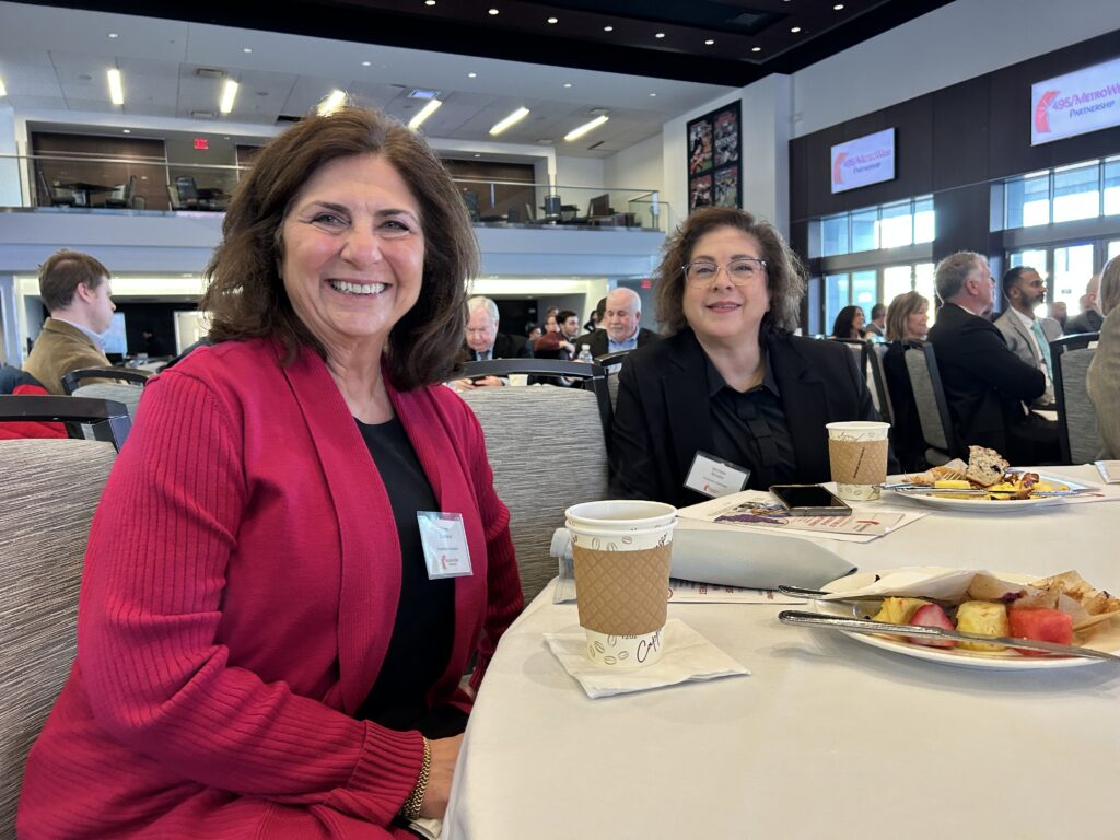 Two smiling women sit at a table in a crowded room.