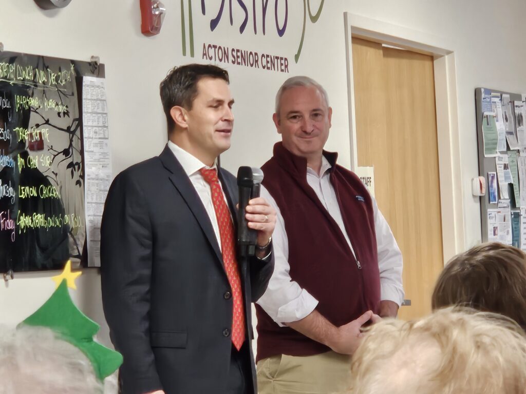 Two men stand at the front of the Acton Senior Center Bistro. One wears a suti and red tie, the other is more casually dressed in a button down shirt and fleece vest.