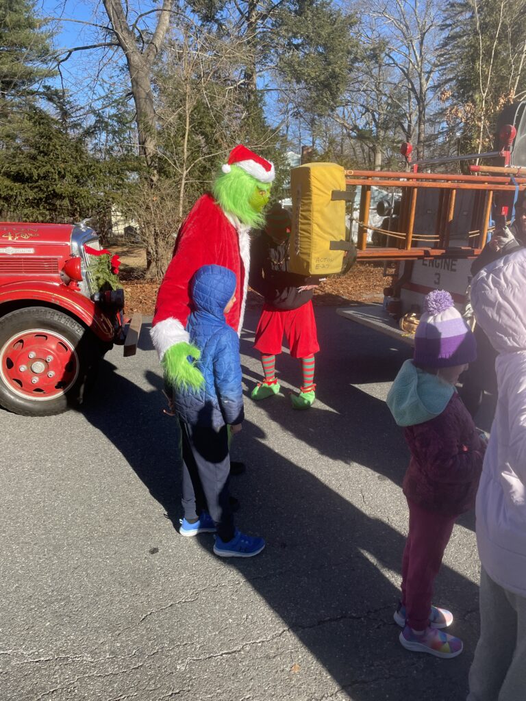 A kid in a blue parka stands with a very tall, skinny, person wearing a Grinch costume (and Santa jacket!). On the far right, you can see a dad's camera pointed at the pair.