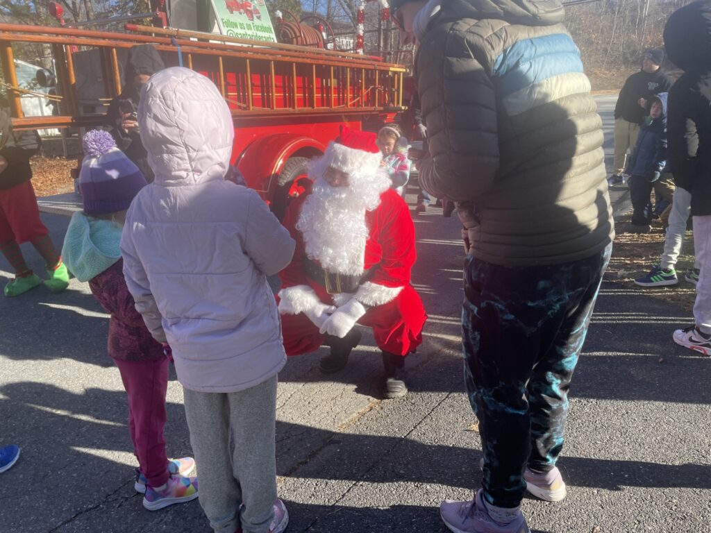 Santa squats down to kid level to talk to two girls in parkas, and a ladder truck is in the background.