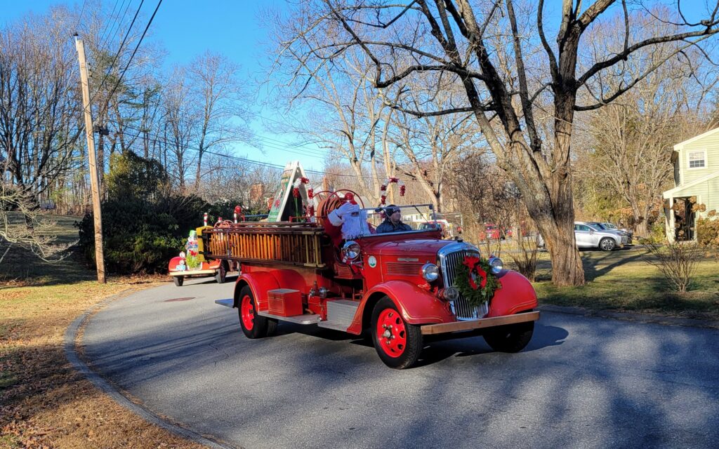 A bright red antique firetruck drives down the street. Santa is in the front seat waving and there are large candy canes on the back of the truck.