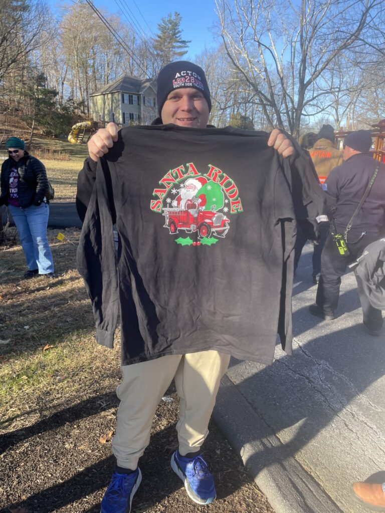 A firefighter wearing a Ladder 28 watch cap holds up a black, long-sleeved, Santa Ride t-shirt.