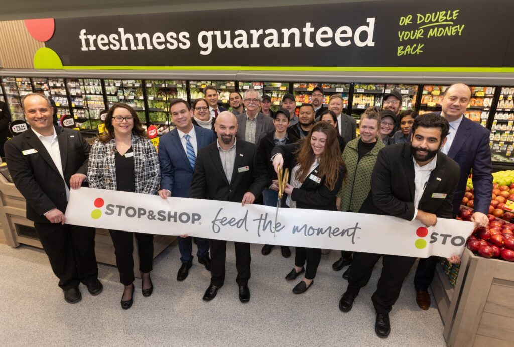 A group of people, including Rep. Dan Sena, Sen. Jamie Eldridge, and Select Board members Fran Arsenault, David Martin, and Alissa Nicol, stand in front of a large ribbon that reads " Stop & Shop. Feed the moment. A woman with oversized scissors is cutting the ribbon.