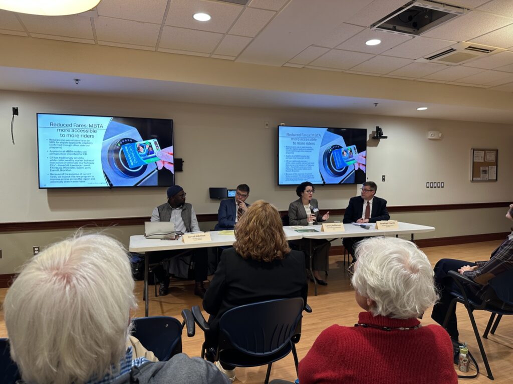 Four people sit at tables in the front of a room. There are two large screens above their heads.