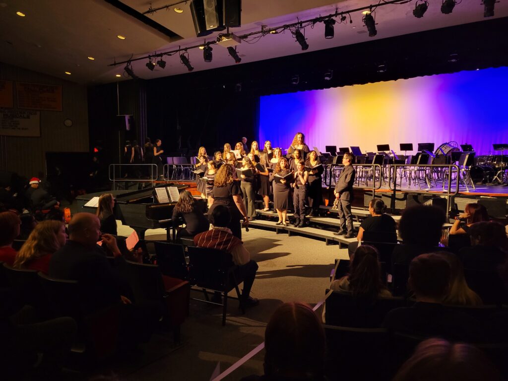 A group of about 20 young women, all wearing black, stand on the risers in front of the stage. They are holding large music folders.