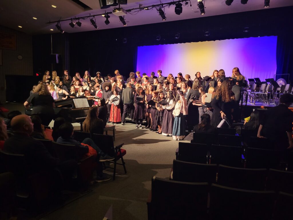 A large group of singers, including the Madrigal singers (in Renaissance garb) stands on risers in front ifthe stage. A piano player is in the center of the photo.