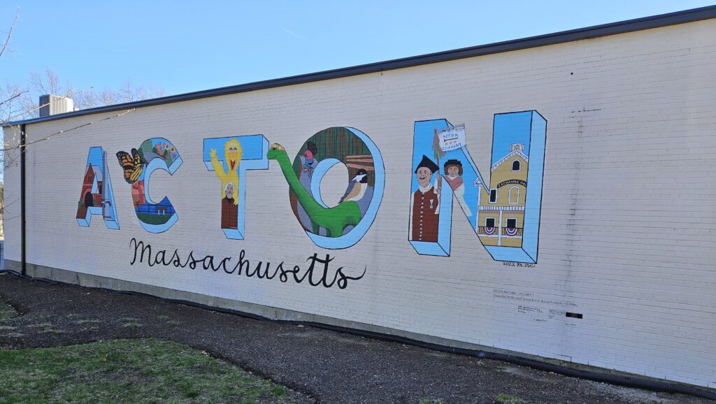 Mural on a white wall that says Acton Massachusetts. The letters in Acton include images associated with Acton such as Bessie the dinosaur, a minuteman, and Big Bird.