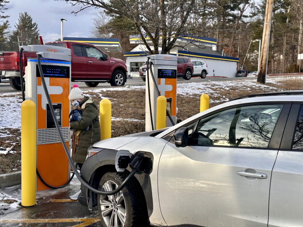 A white car is pluged into the large orange Chargepoint charger. A woman stands next to the charger.