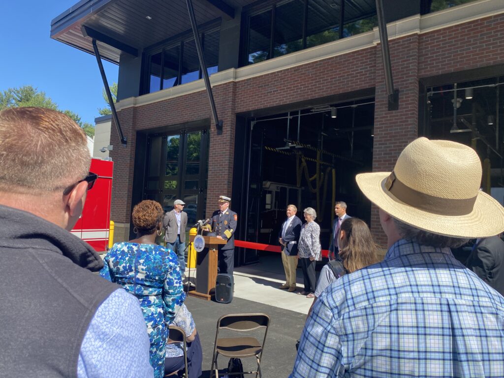 People standing in front of the North Acton fire station. The fire chief at the time is giving a speech at a podium and a red ribbon is across one of the open bay doors.